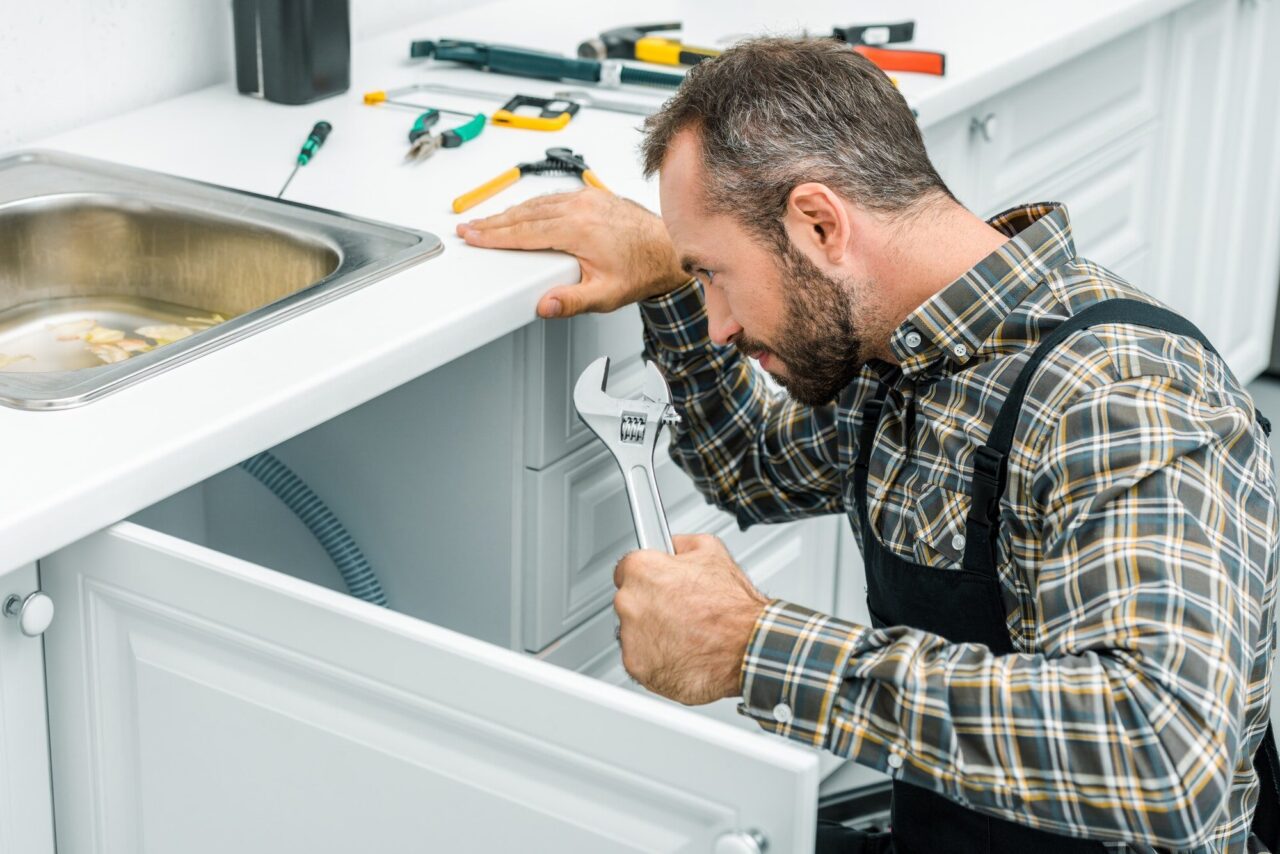 A man in plaid shirt and overalls kneels by a white kitchen cabinet with a wrench. Various tools are on the counter. The mood is focused and diligent.