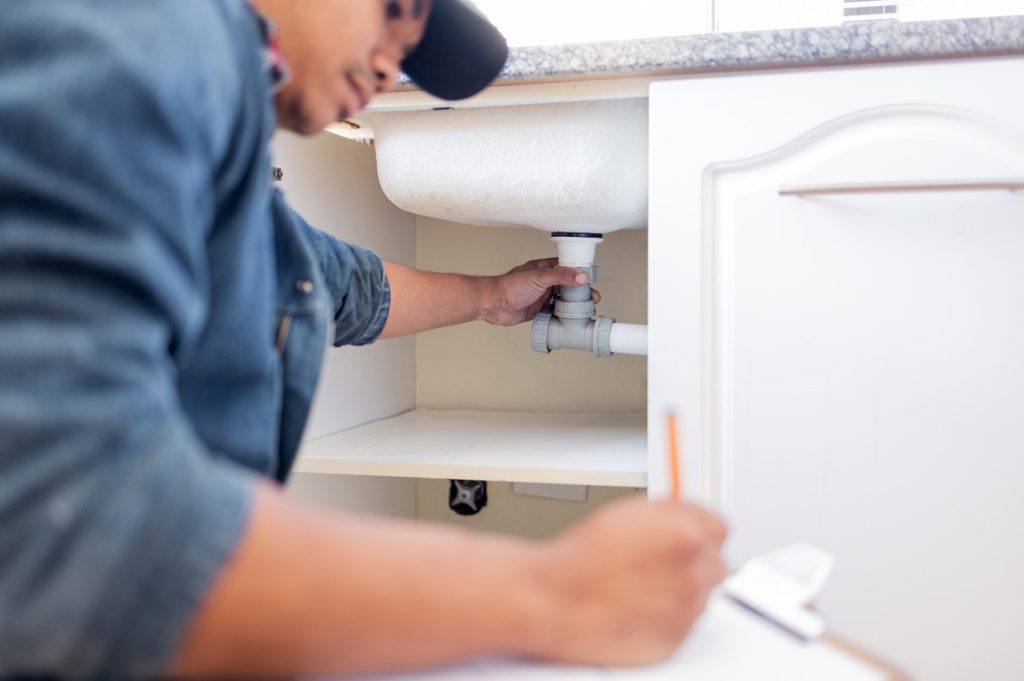 Plumber performing plumbing services under a kitchen sink to check for leaks and loose pipe connections.