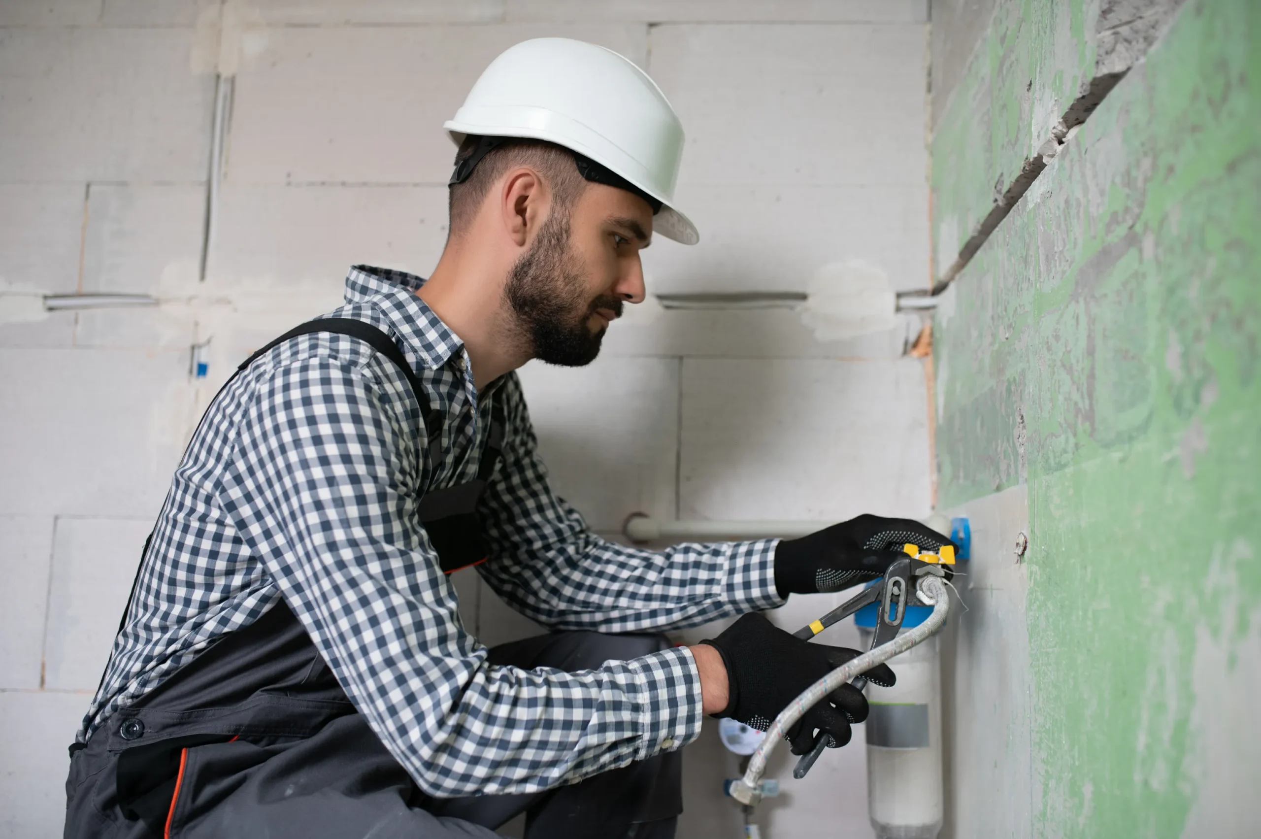 A construction worker in a hard hat and gloves installs a wall fixture, focused and determined. The setting is a partially finished room with green drywall.